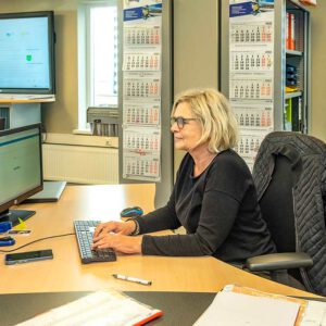 een vrouw met blond haar en een bril zit achter een bureau in een kantoor en typt op een toetsenbord