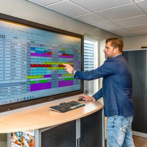 Een man in een colbert en spijkerbroek wijst naar een groot interactief scherm waarop een rooster of planning staat, in een moderne kantooromgeving.