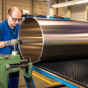 Een fabrieksmedewerker met een bril en werkhandschoenen in een blauwe overall is bezig met het bewerken of rollen van een groot, glanzend metalen cilinder op een machine in een werkplaats.