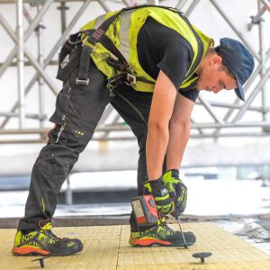Een bouwvakker met veiligheidsharnas, helm en werkhandschoenen is bezig met het vastzetten van isolatiemateriaal (Rockwool) op een dak met behulp van een boormachine.