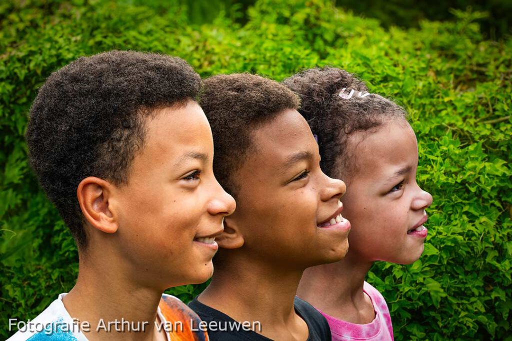 portretfotografie. Drie lachende kinderen in profiel staan naast elkaar, met een groene, met klimop begroeide heg als achtergrond.