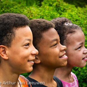portretfotografie. Drie lachende kinderen in profiel staan naast elkaar, met een groene, met klimop begroeide heg als achtergrond.