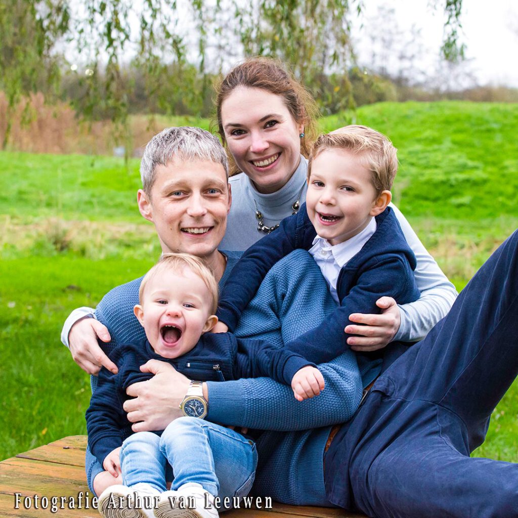 groeps- en familiefotografie. Een lachende familie zit op een houten bank. De man heeft een baby op schoot en de vrouw zit dicht tegen hem aan, met hun oudere kind op haar schoot. Ze kijken allemaal vrolijk in de camera.