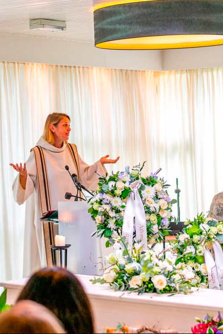 Uitvaartfoto van een voorgangster in een witte stola die spreekt bij de kist met bloemstukken.