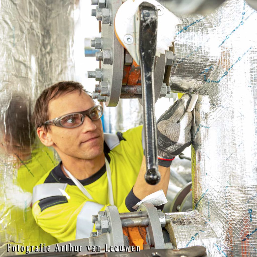 Een werknemer met veiligheidsbril en handschoenen isoleert een buis. Hij gebruikt Rockwool isolatiemateriaal.