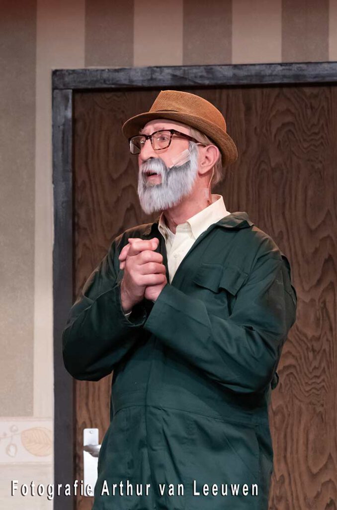 Theaterfoto van een acteur met hoed, bril en een nepbaard in een groen overall.