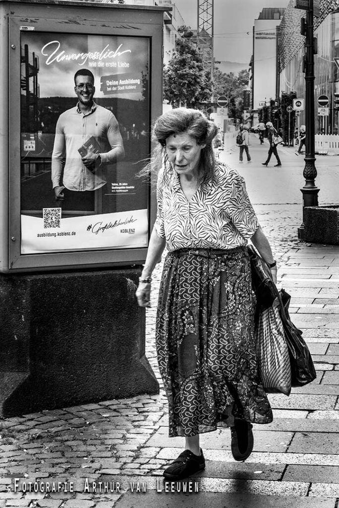 Zwart-wit straatfoto van een oudere vrouw die met boodschappentassen over de stoep loopt, langs een reclamebord.