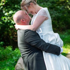 Bruidspaar omhelst elkaar, man tilt vrouw op in trouwjurk, buiten in de natuur.