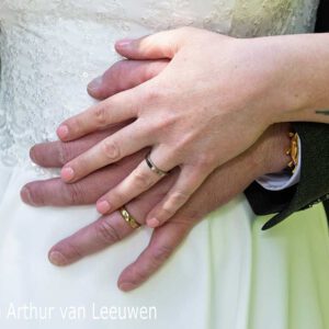 Close-up van handen van het bruidspaar op de witte trouwjurk, met trouwringen zichtbaar.