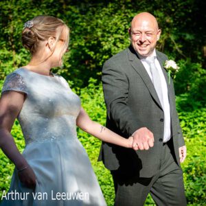 Bruidspaar loopt hand in hand door groene natuur, beiden lachend.