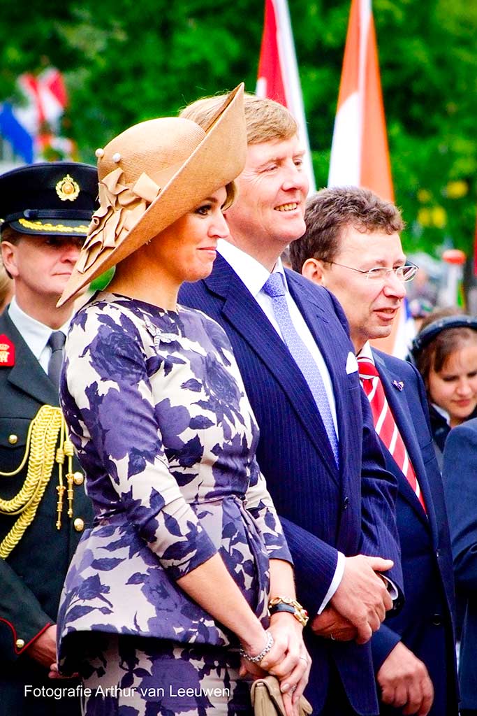 Prinses Máxima en Prins Willem-Alexander (nu Koning en Koningin) tijdens nationale feestdag of evenement, omgeven door Nederlandse vlaggen en militaire/burgerfunctionarissen.