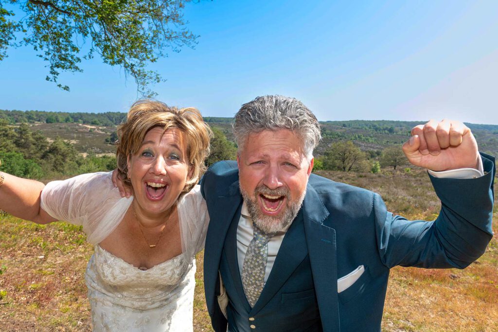 Een enthousiast en lachend bruidspaar viert feest in de natuur met een wijds uitzicht over de heide en bossen op de achtergrond.