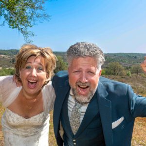 Een enthousiast en lachend bruidspaar viert feest in de natuur met een wijds uitzicht over de heide en bossen op de achtergrond.