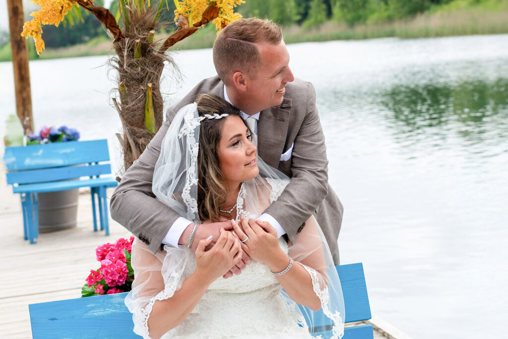 Romantisch bruidspaar zittend op een blauwe bank aan het water, waarbij de bruidegom de bruid van achteren omhelst tijdens een fotoshoot op hun trouwdag. fotografie arthur van leeuwen