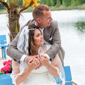 Romantisch bruidspaar zittend op een blauwe bank aan het water, waarbij de bruidegom de bruid van achteren omhelst tijdens een fotoshoot op hun trouwdag. fotografie arthur van leeuwen