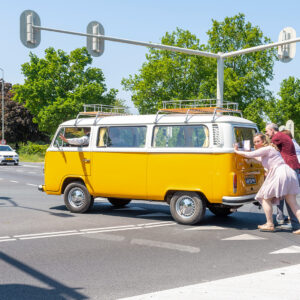 Gasten in feestkleding duwen een gele klassieke Volkswagen T2 trouw bus vooruit op een kruispunt tijdens een bruiloft.
