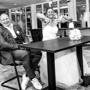 Zwart-wit foto van een lachend bruidspaar aan een tafel tijdens de ceremonie, waarbij de bruid een grappige pose maakt en de bruidegom sneakers onder zijn trouwpak draagt.