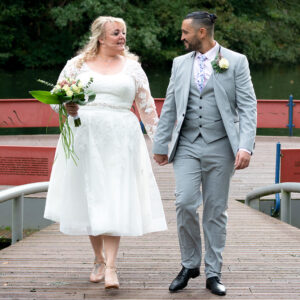 Een bruidspaar loopt hand in hand over een houten brug aan het water, waarbij de bruid een korte kanten trouwjurk draagt en de bruidegom een lichtgrijs trouwpak. sonsbeek-arnheh-fotografie arthur van leeuwen