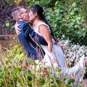 Romantisch bruidspaar kust elkaar innig tijdens hun trouwreportage in een bloemrijke tuin, met de bruidegom in een blauw pak en de bruid in een kanten trouwjurk. Nijmegen_fotografie arthur van leeuwen