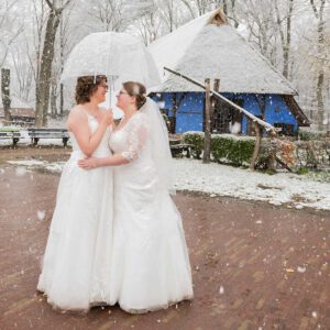 Twee bruiden in witte trouwjurken omhelzen elkaar onder een transparante paraplu tijdens een winterbruiloft in de sneeuw bij een blauwe historische boerderij.
