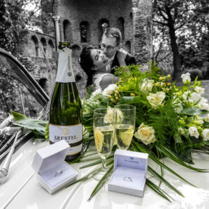 Zwart-wit trouwfoto van trouwringen in doosjes, een fles Arestel cava en een bruidsboeket op de motorkap van een trouwauto, met een kussend bruidspaar bij een ruïne op de achtergrond.